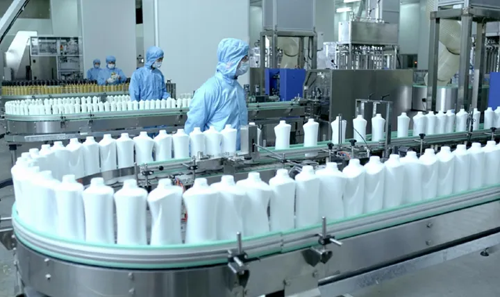 Workers in blue uniforms and hairnets oversee a factory production line with numerous white bottles on a conveyor belt.